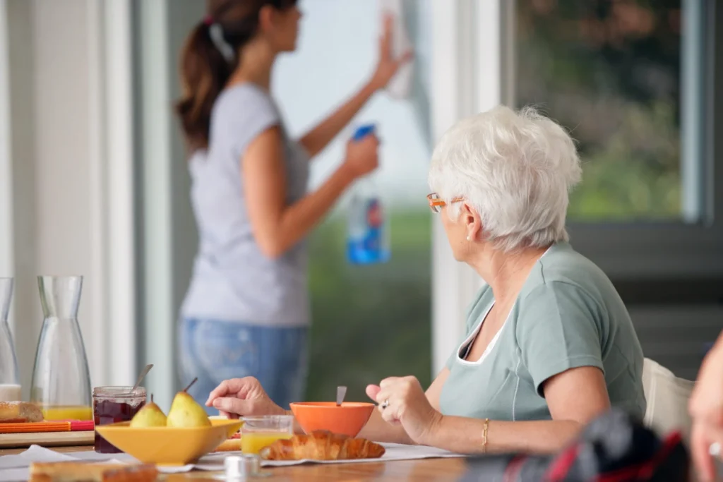Aide ménagère nettoyant la cuisine pendant qu’une femme âgée prend son petit-déjeuner