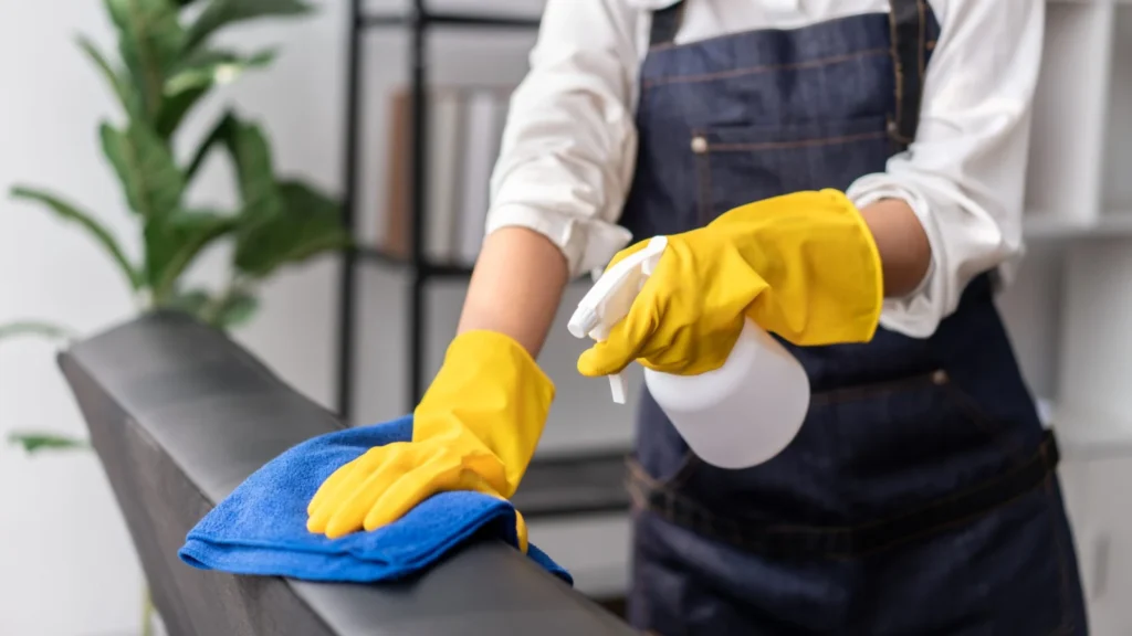 Aide ménagère vaporisant et essuyant un meuble avec des gants jaunes