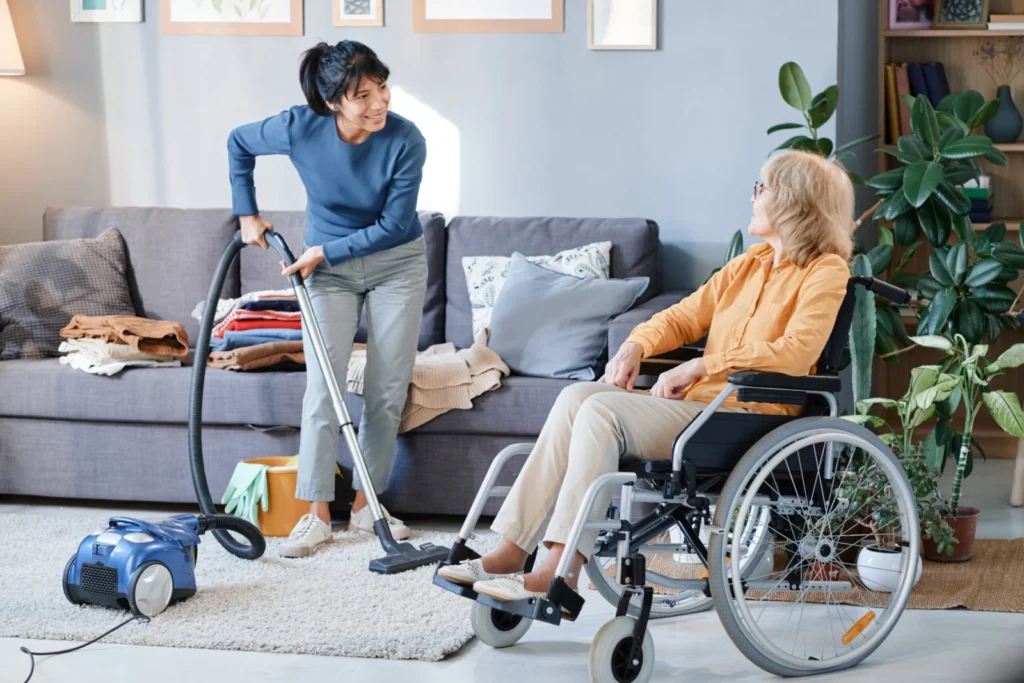 Aide ménagère passant l’aspirateur près d’une femme en fauteuil roulant